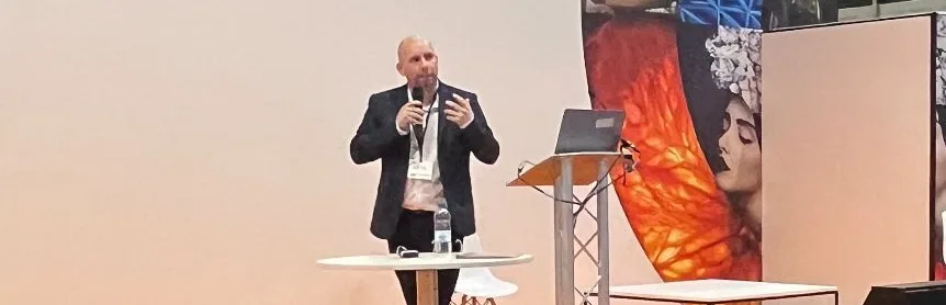 Lee Brooker on stage at a photography conference, holding a microphone mid-sentence.